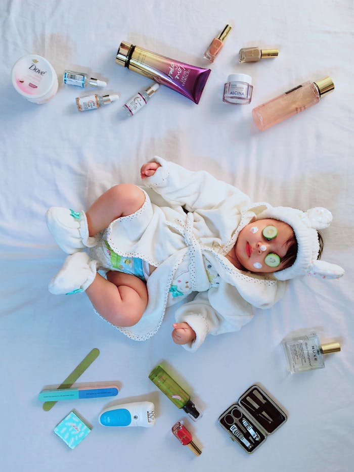 Cute baby with cucumber eye mask surrounded by skincare items on a bed.